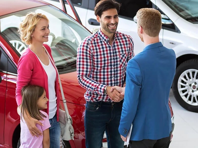 A family talking with a dealership representative while standing next to a red vehicle